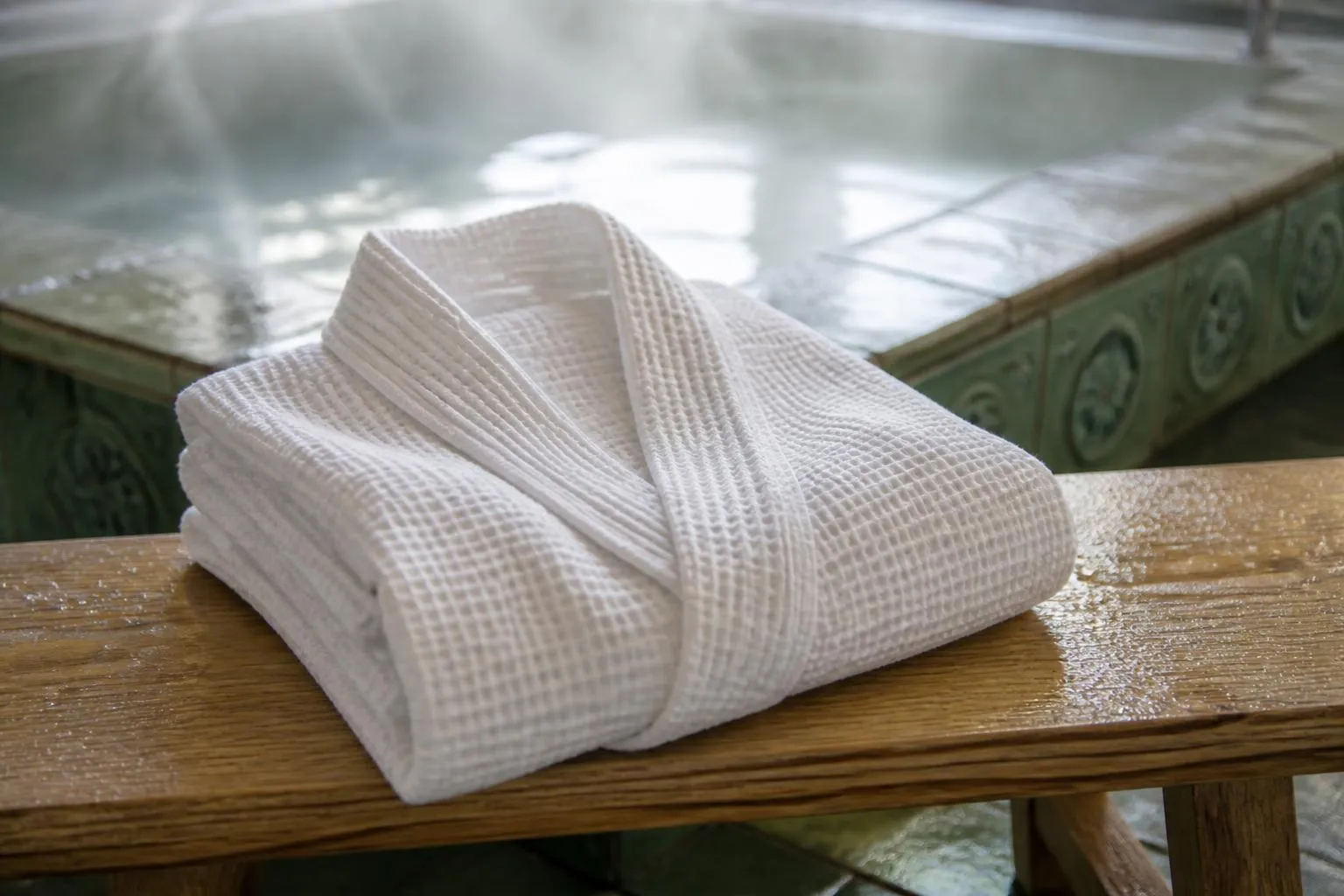 Un peignoir blanc plié sur un banc de bois clair, au bord d'un bassin thermal dont l'eau fume légèrement.