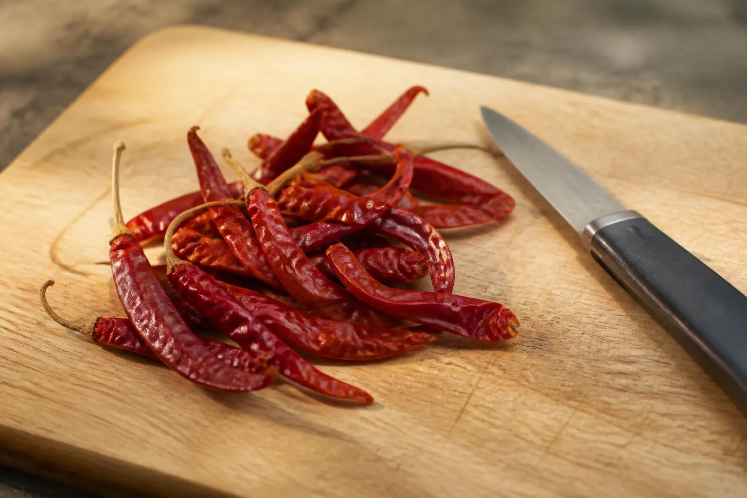 Des piments rouges séchés posés sur une planche à découper en bois, à côté d'un couteau d'office