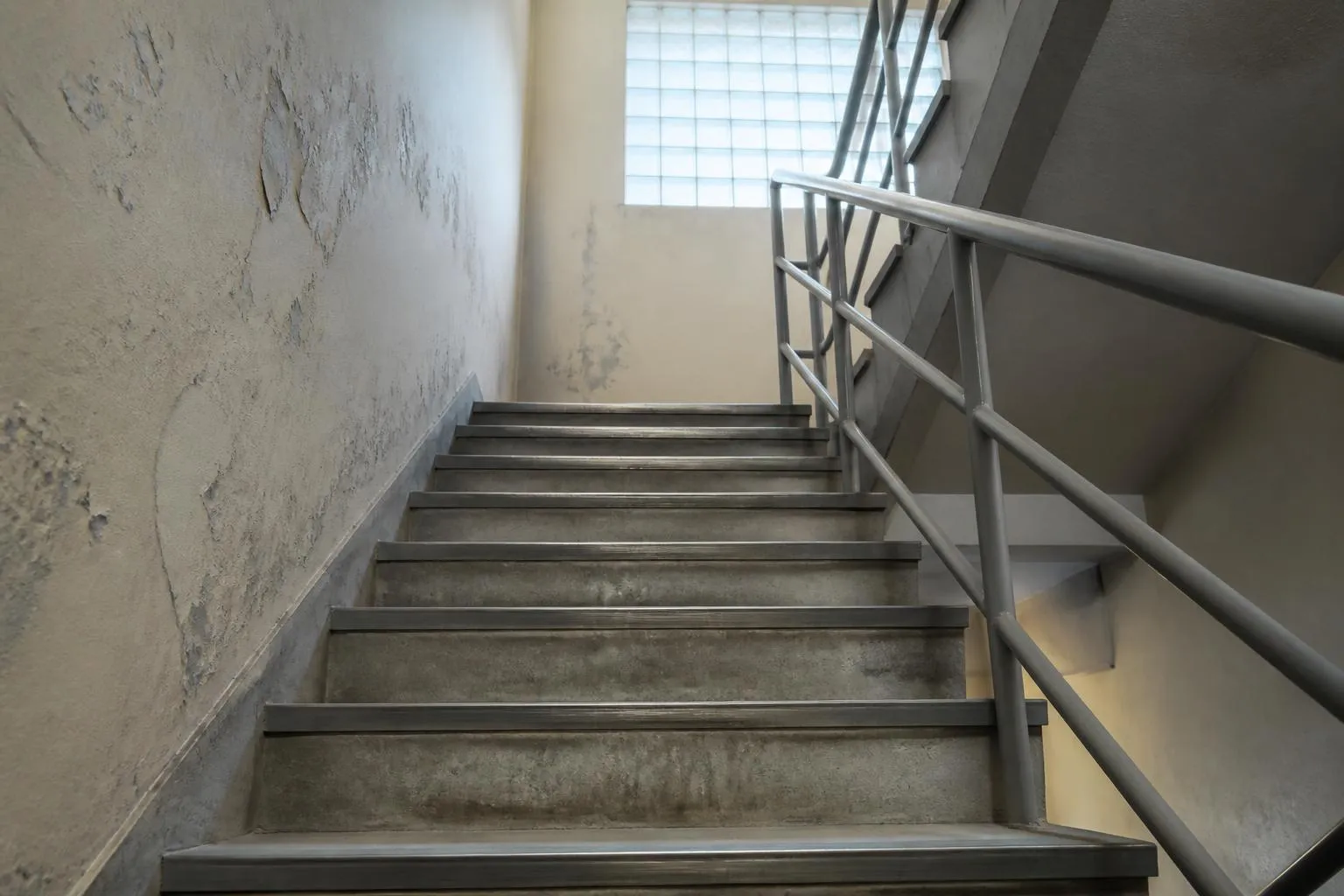 Cage d'escalier de service en béton peint, rampe métallique, marches vues depuis le bas